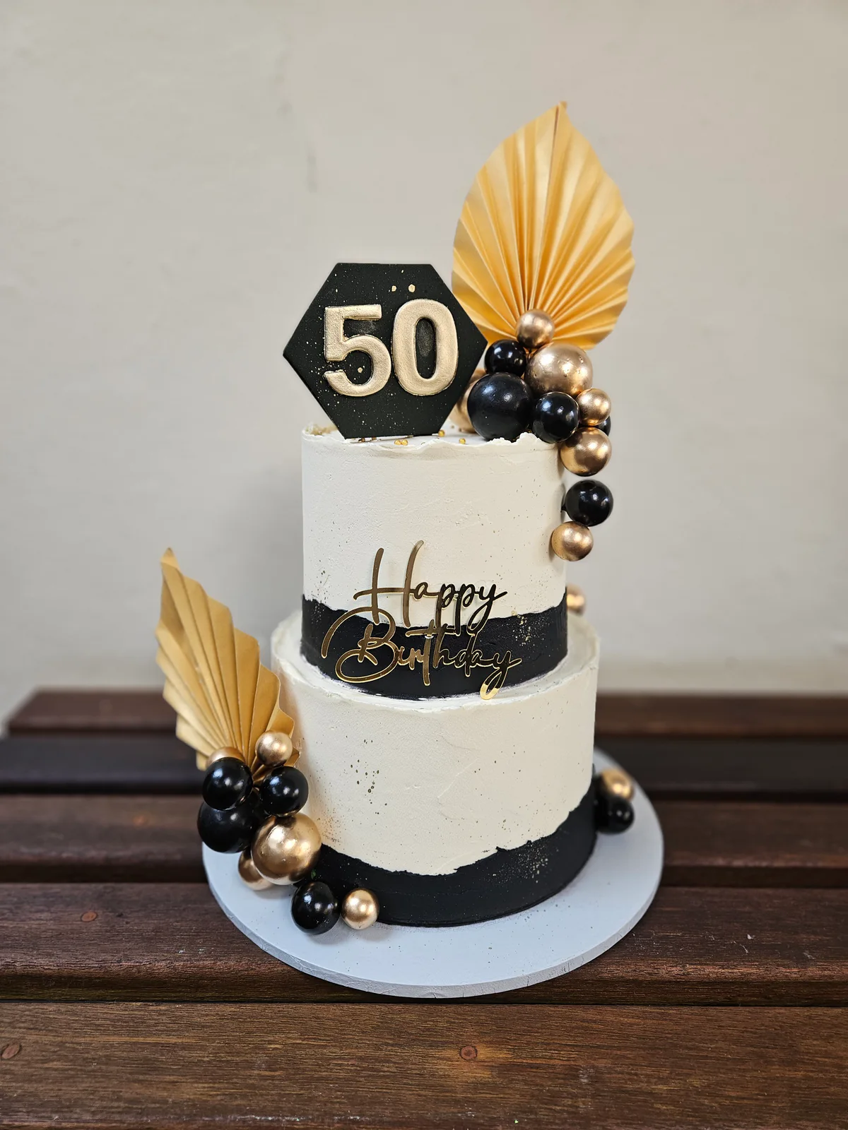 Two-tier white fiftieth birthday cake with black and gold accents, fan toppers and decorative spheres.