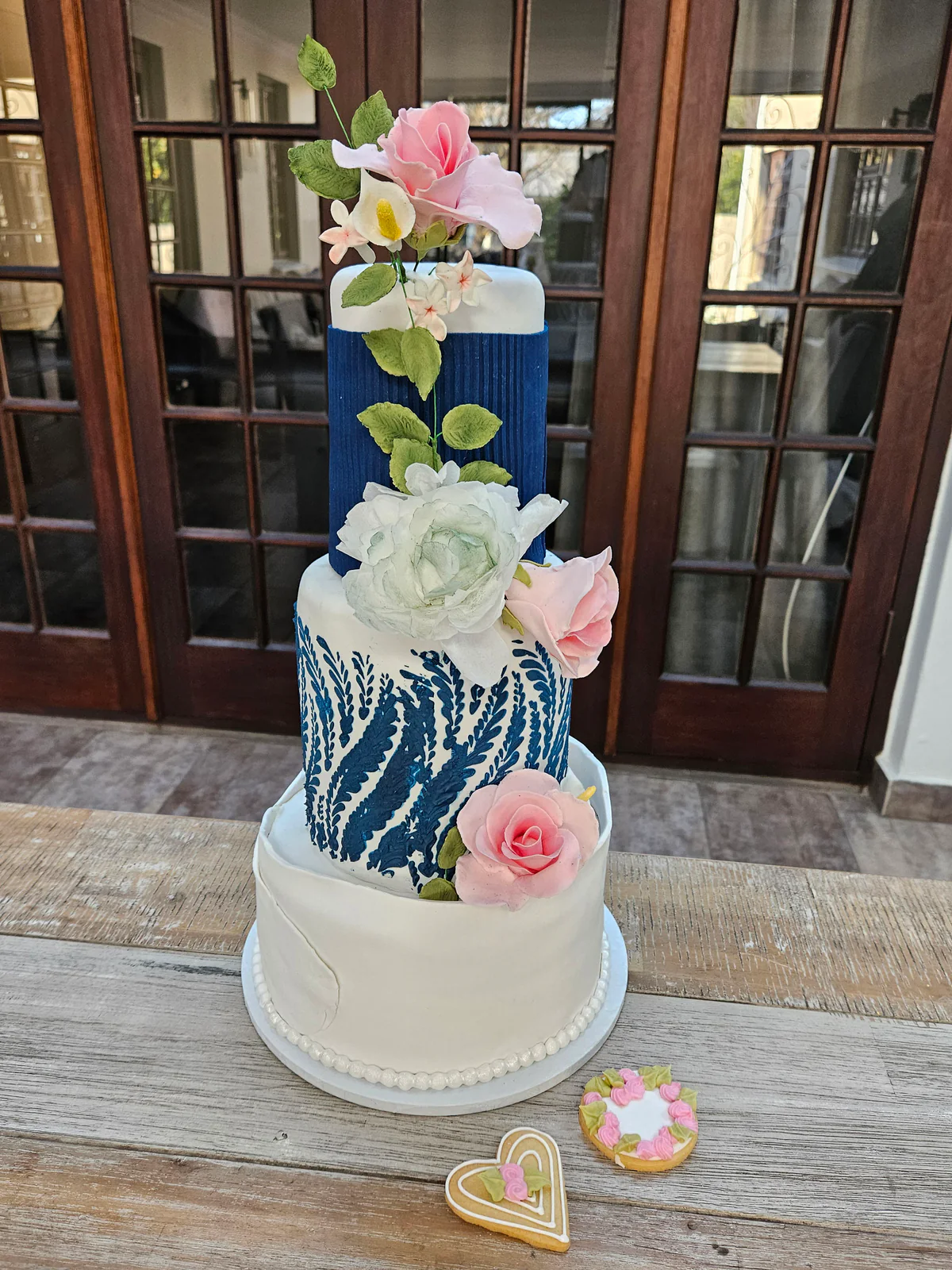 Blue and white tiered class cake with textured patterns, pink and white flowers and decorated biscuits beside it.