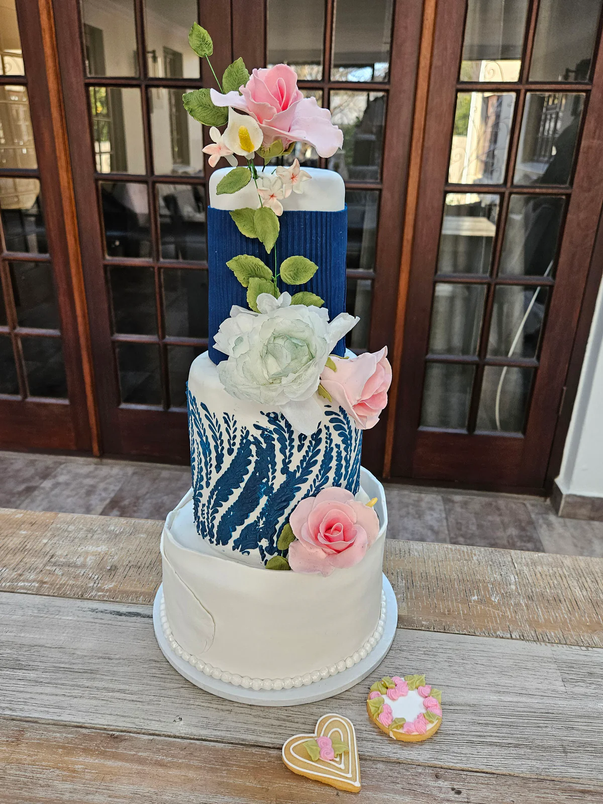 Blue and white tiered class cake with textured patterns, pink and white flowers and decorated biscuits beside it.