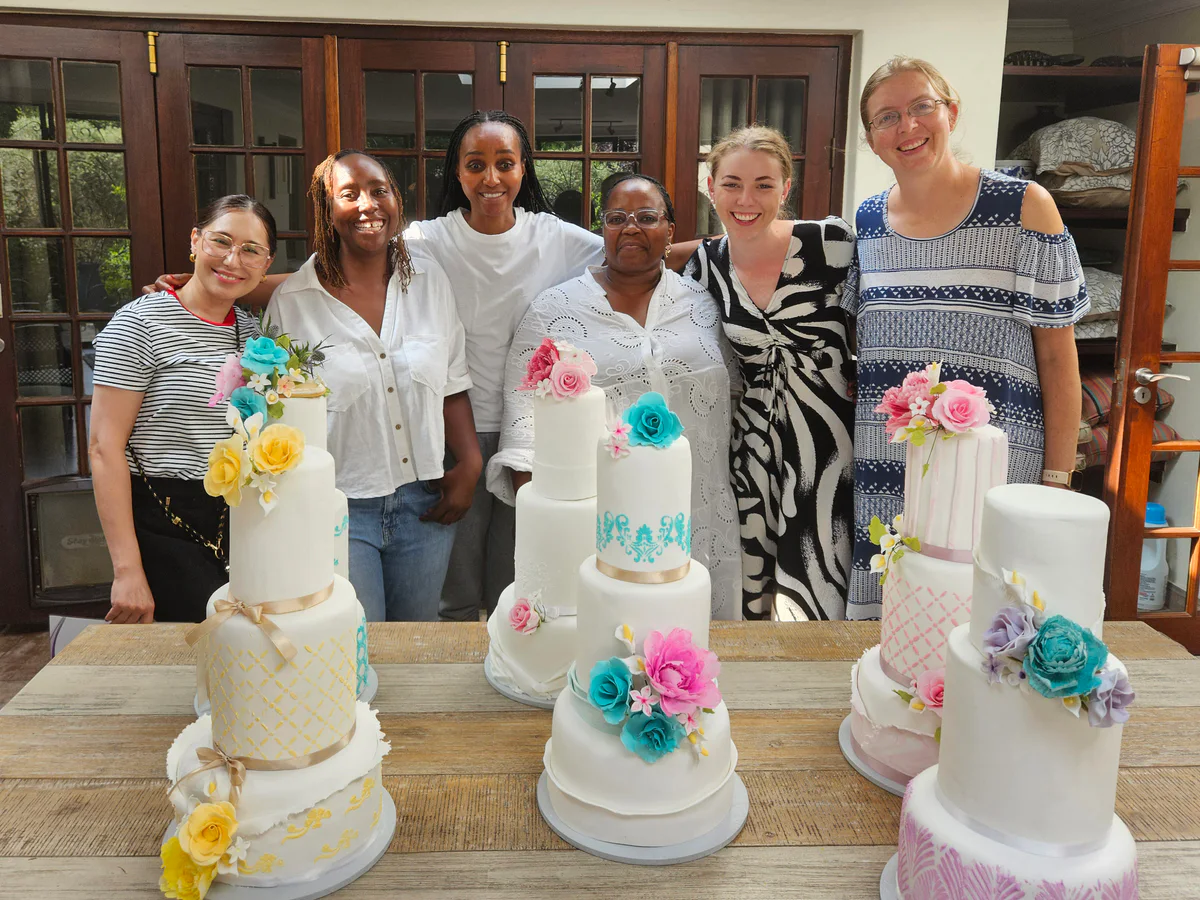 Cake decorating class group standing behind a display of finished tiered cakes with colourful floral decorations.