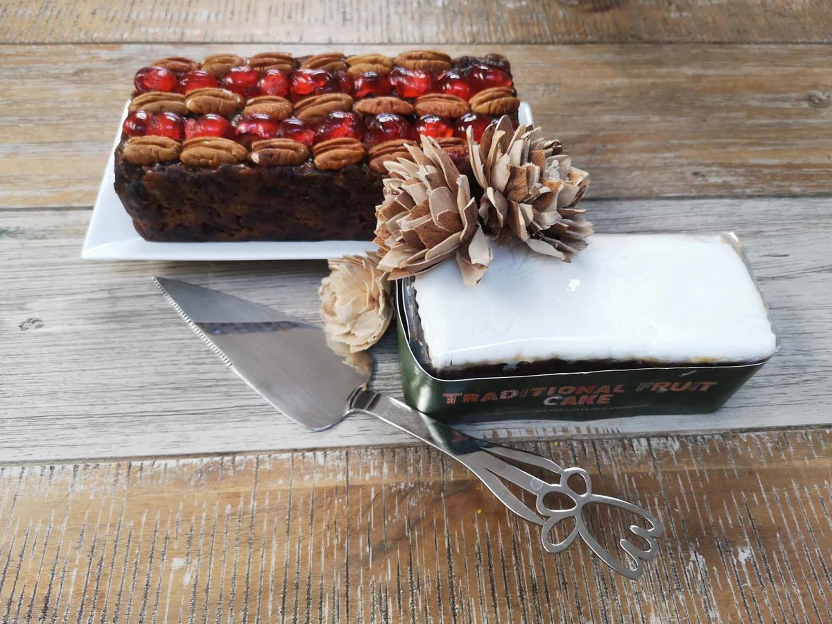 Two rectangular fruit cakes, one with white icing and pine-cone decorations and one topped with nuts and cherries.