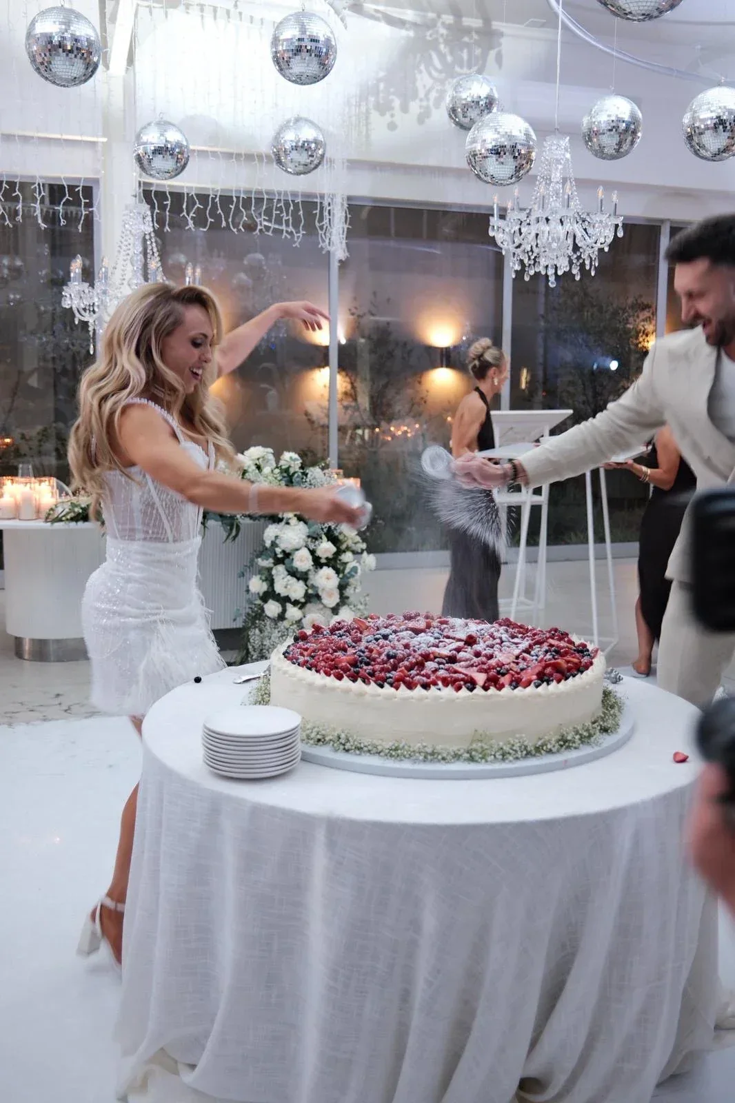 Bride and groom dusting icing sugar over a large berry-topped wedding cake beneath chandeliers and mirrored disco balls.
