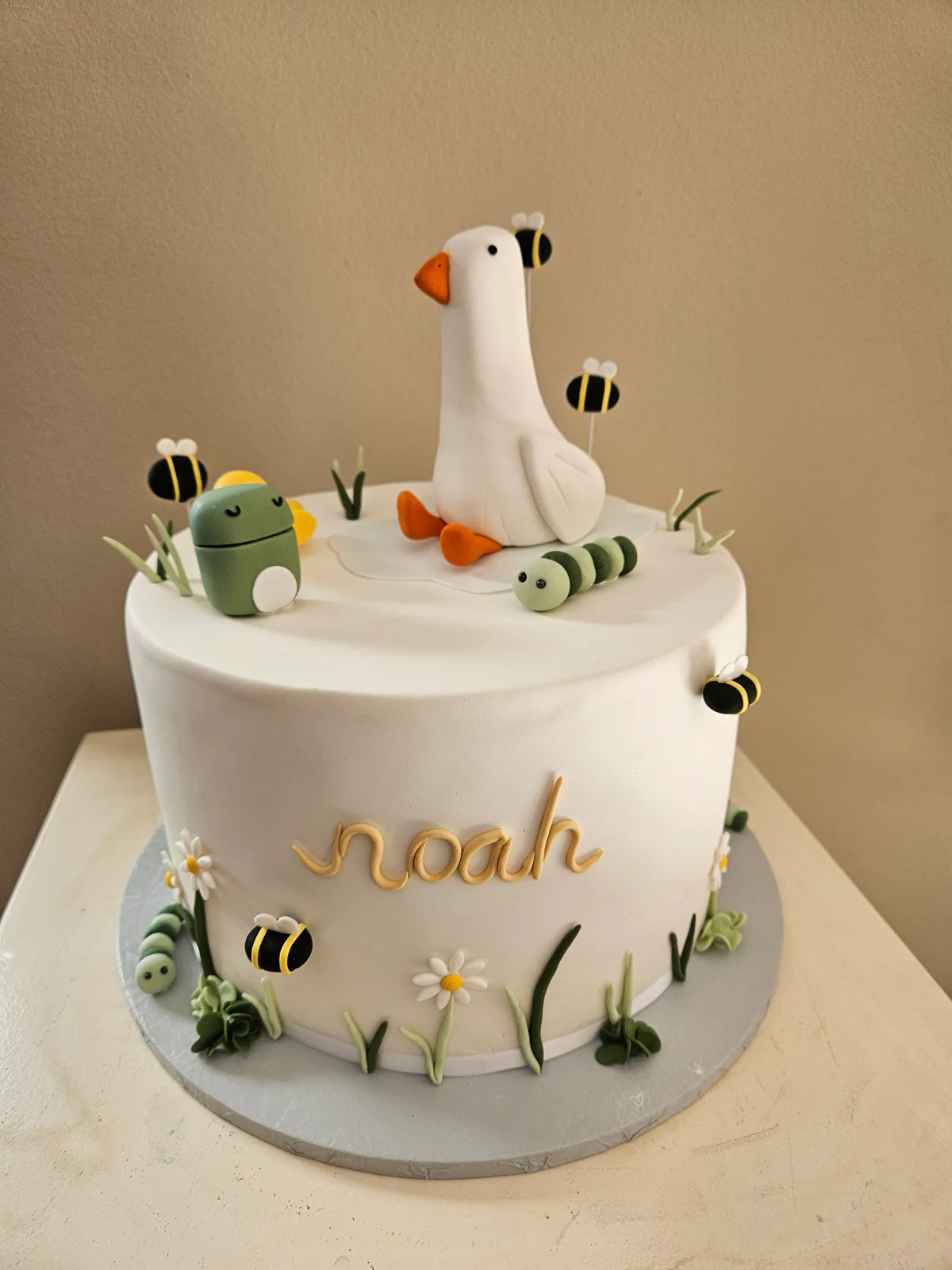 White first birthday cake with a goose-shaped number one, bees, grass and a small green watering can.