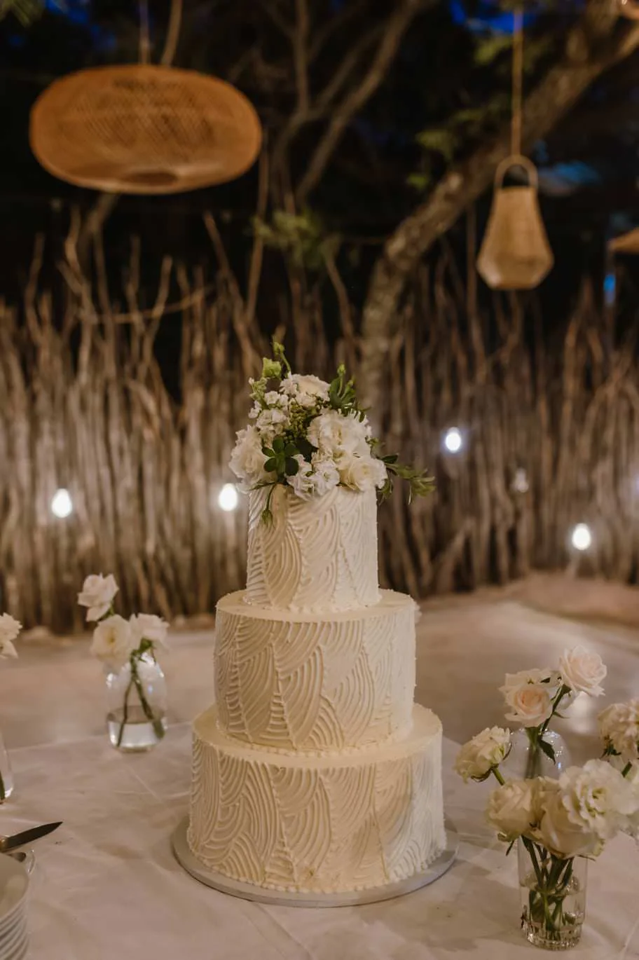Three-tier ivory textured wedding cake topped with white flowers at an evening reception.