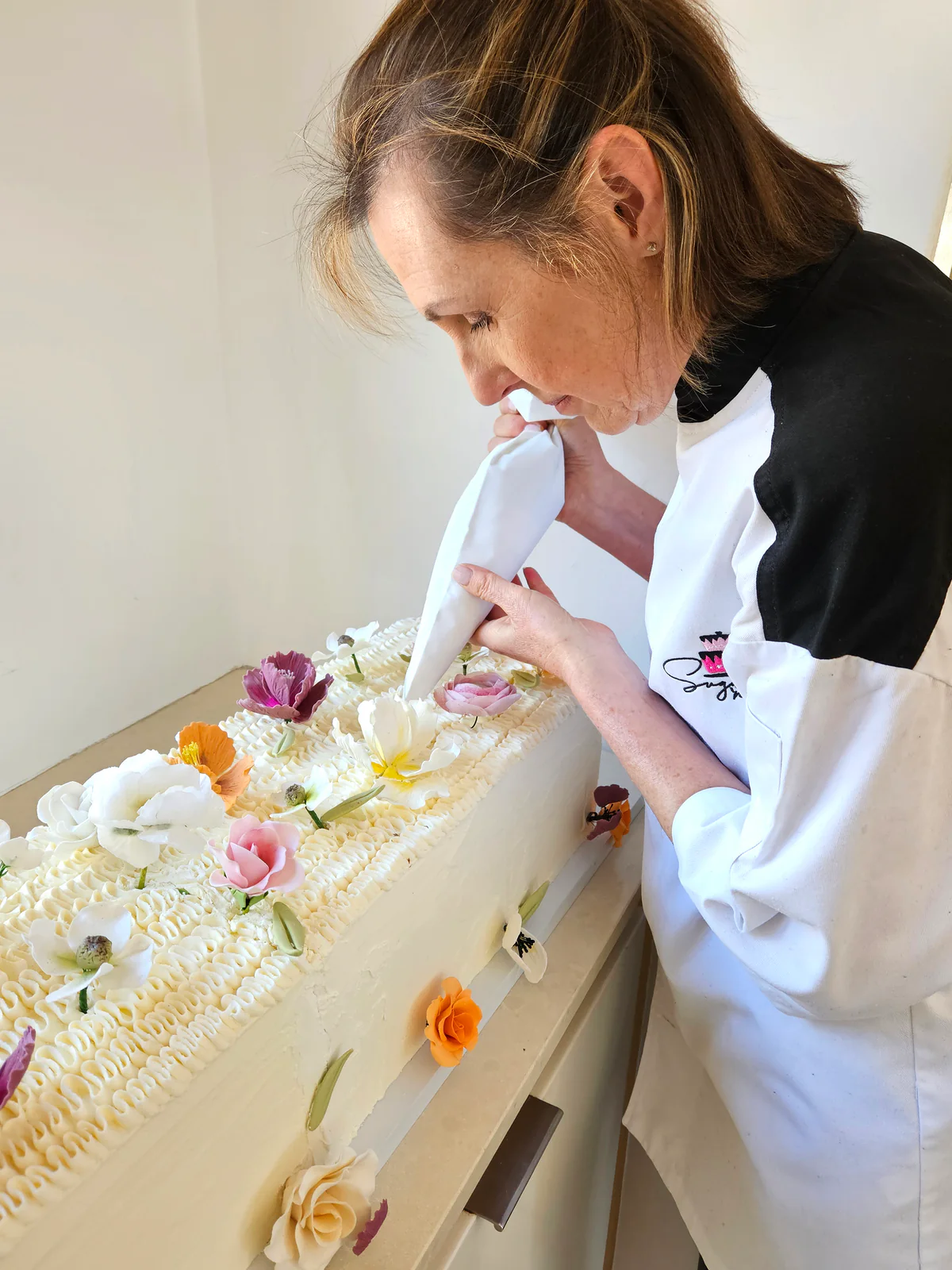 Kate piping white icing onto a long rectangular cake decorated with pink, purple, yellow and white flowers.
