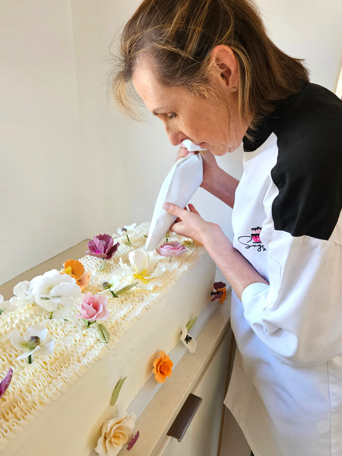 Kate piping white icing onto a long rectangular cake decorated with pink, purple, yellow and white flowers.