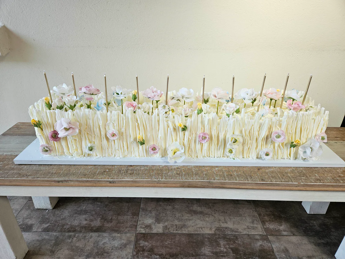Long rectangular white wedding cake with tall piped strands and scattered pastel flowers.