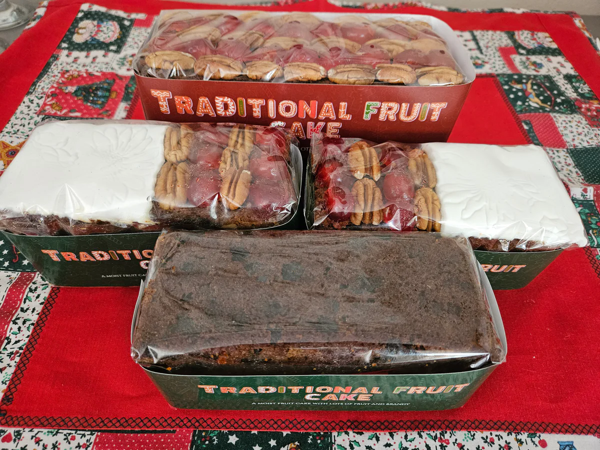 Plain rectangular Christmas fruit cake in festive packaging, with iced and pecan-and-cherry-topped fruit cakes behind it.