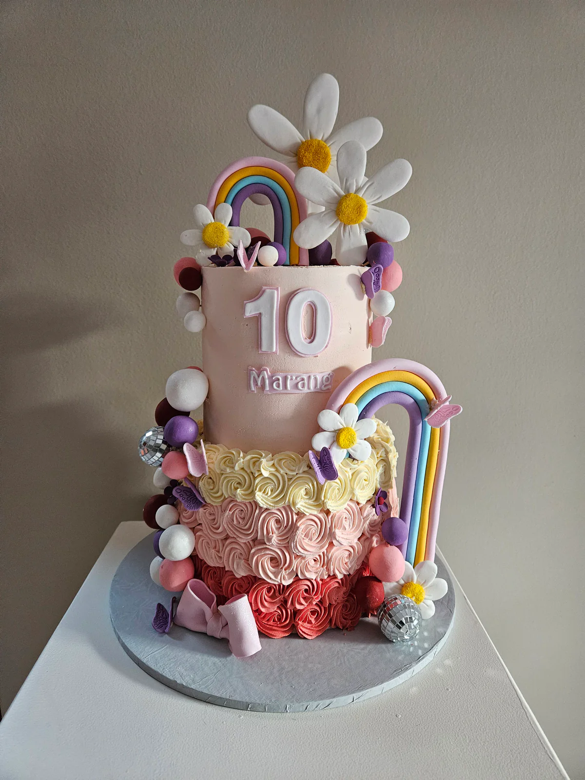 Two-tier pink tenth birthday cake with rainbows, large daisies, piped roses and pastel spheres.