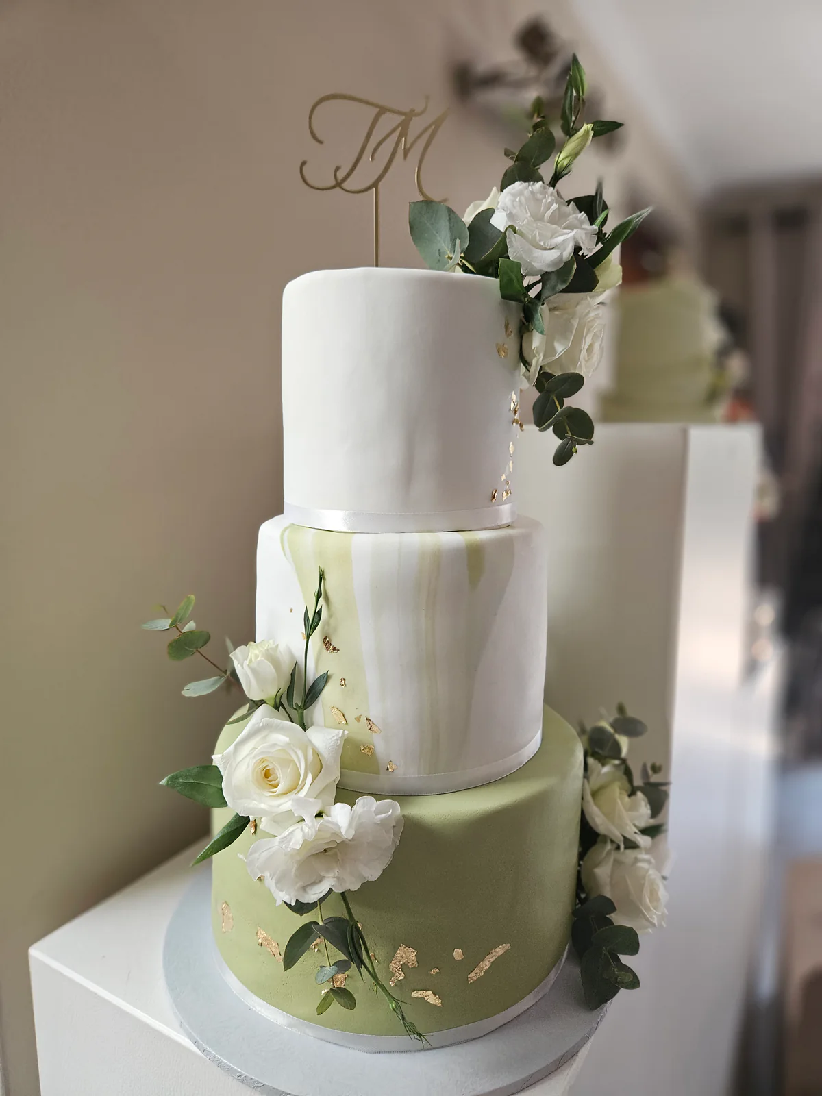 Three-tier sage green and white wedding cake with white roses, green foliage, gold-coloured accents and a monogram topper.