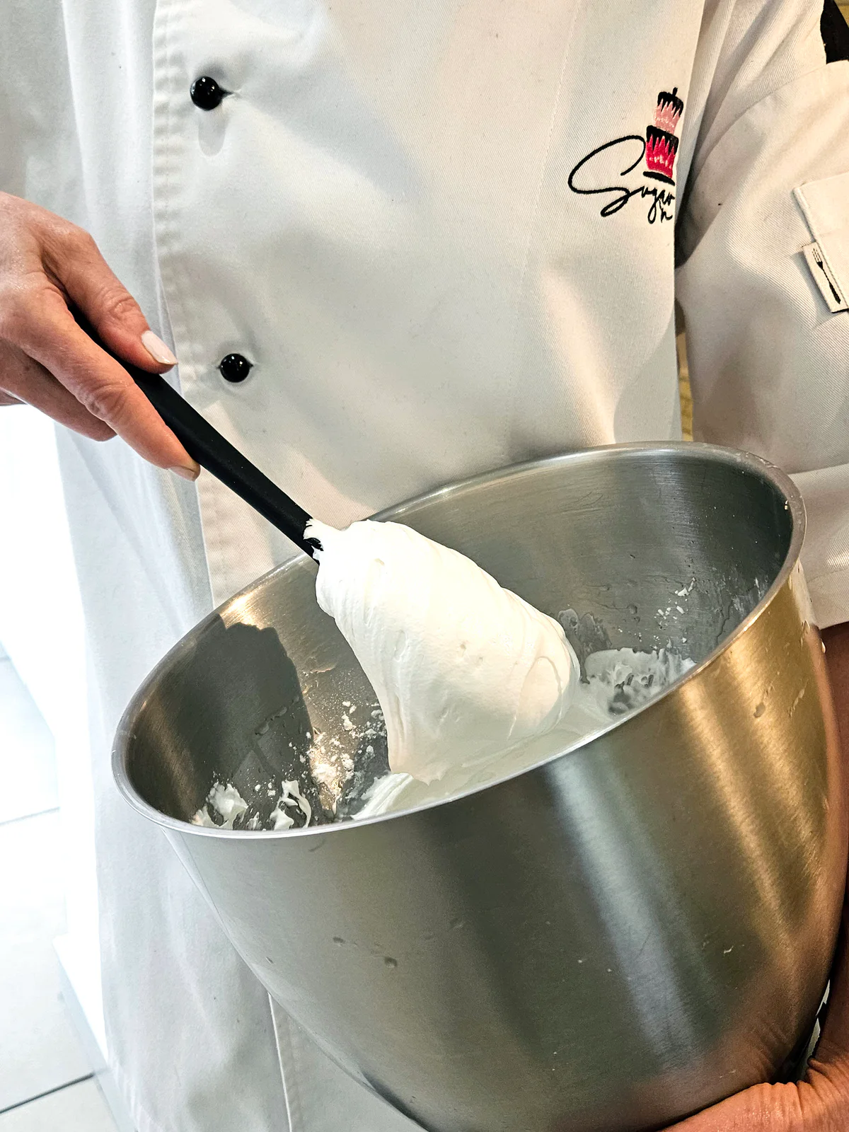Close-up of white icing on a spatula above a stainless steel mixing bowl held by Kate.