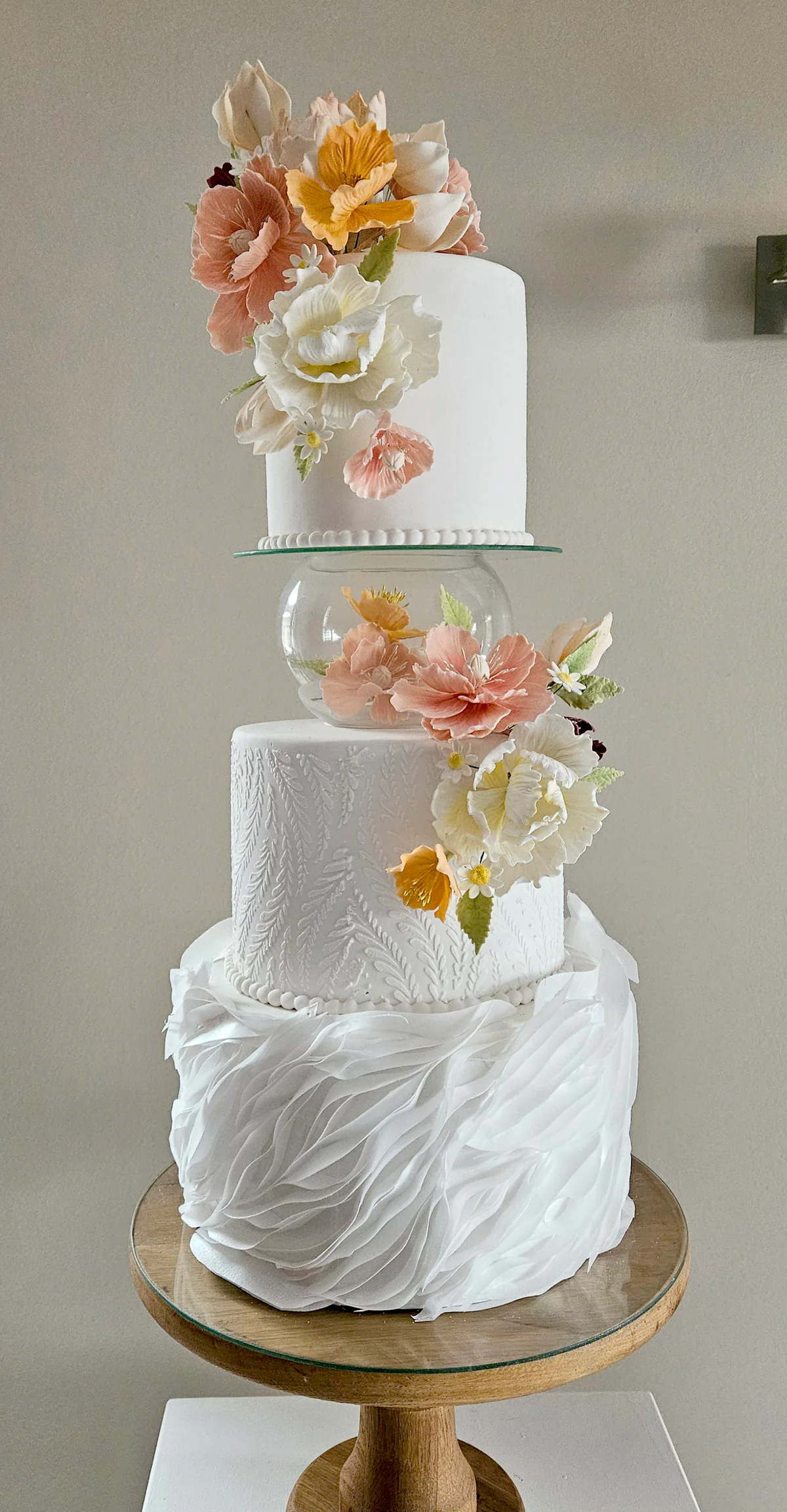 White wedding cake with draped and textured tiers, a clear separator and peach and yellow flowers.