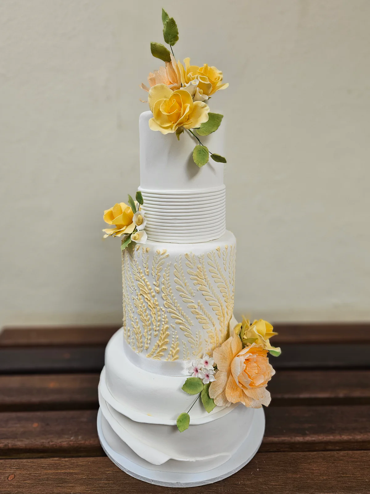 Tall white tiered wedding cake with yellow and peach flowers, fine horizontal ridges and layered curved panels.