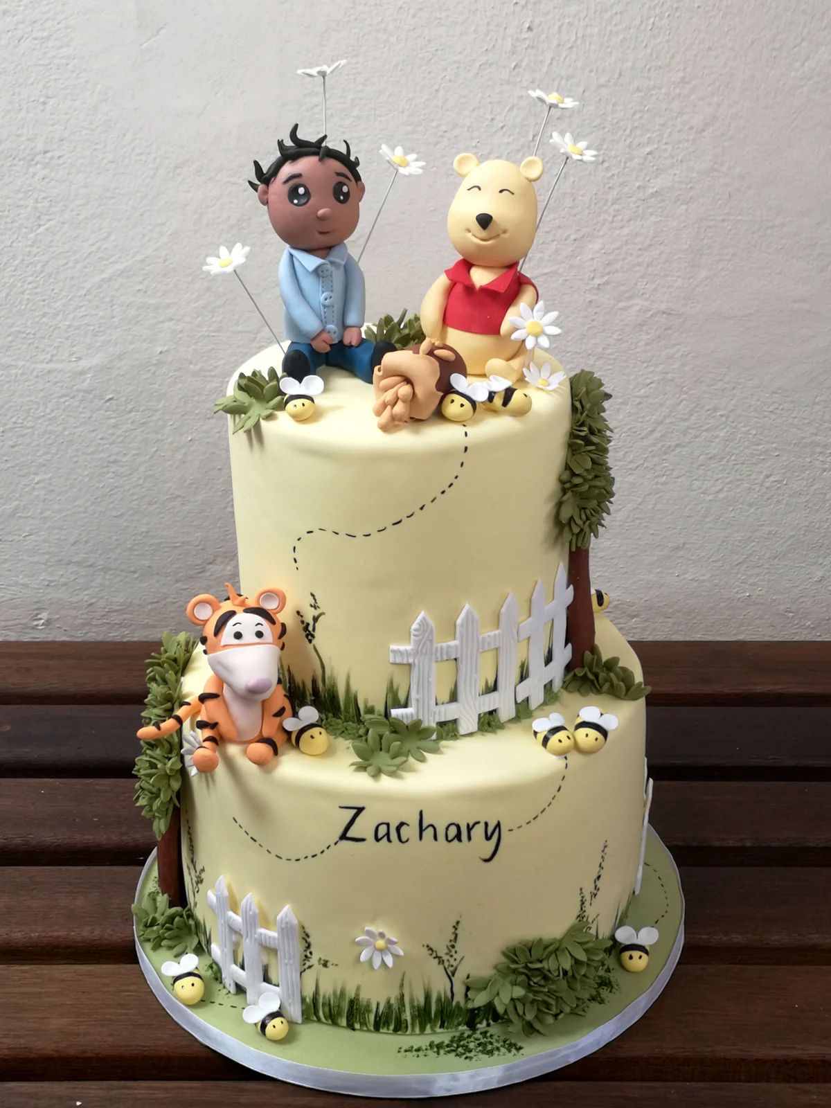Two-tier pale green Winnie the Pooh cake with character figures, daisies and white picket fences.